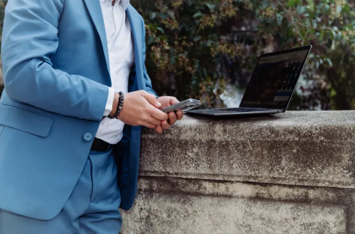 Un homme en costume bleu clair, vu à mi-corps, tient son téléphone près d’un ordinateur portable posé sur une balustrade de pierre.