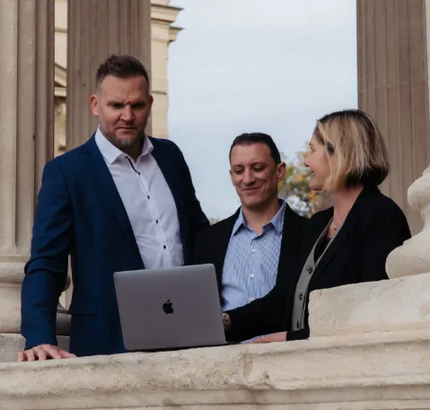 Trois membres de l’équipe de PCC échangent devant un ordinateur portable, au pied d’une colonnade.