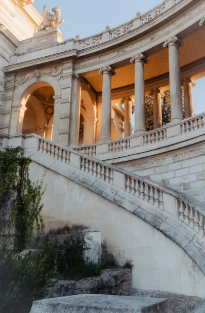 Le grand escalier courbe du Palais Longchamp, sa balustrade de pierre et ses colonnes, dans une lumière dorée.