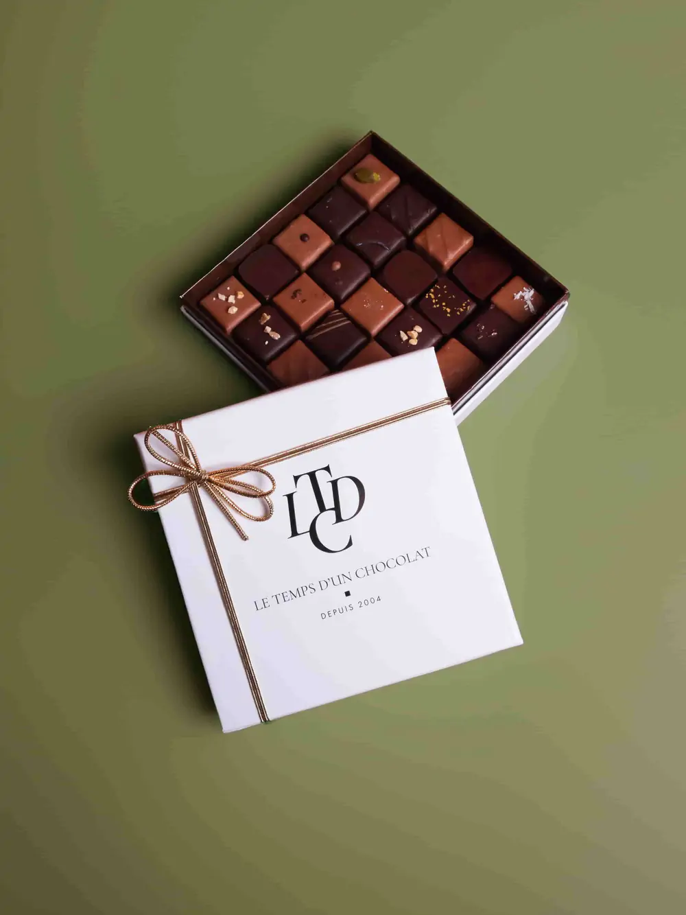 Le Temps d’un Chocolat — un coffret ouvert de chocolats et son coffret blanc noué d’un ruban doré, sur un fond vert