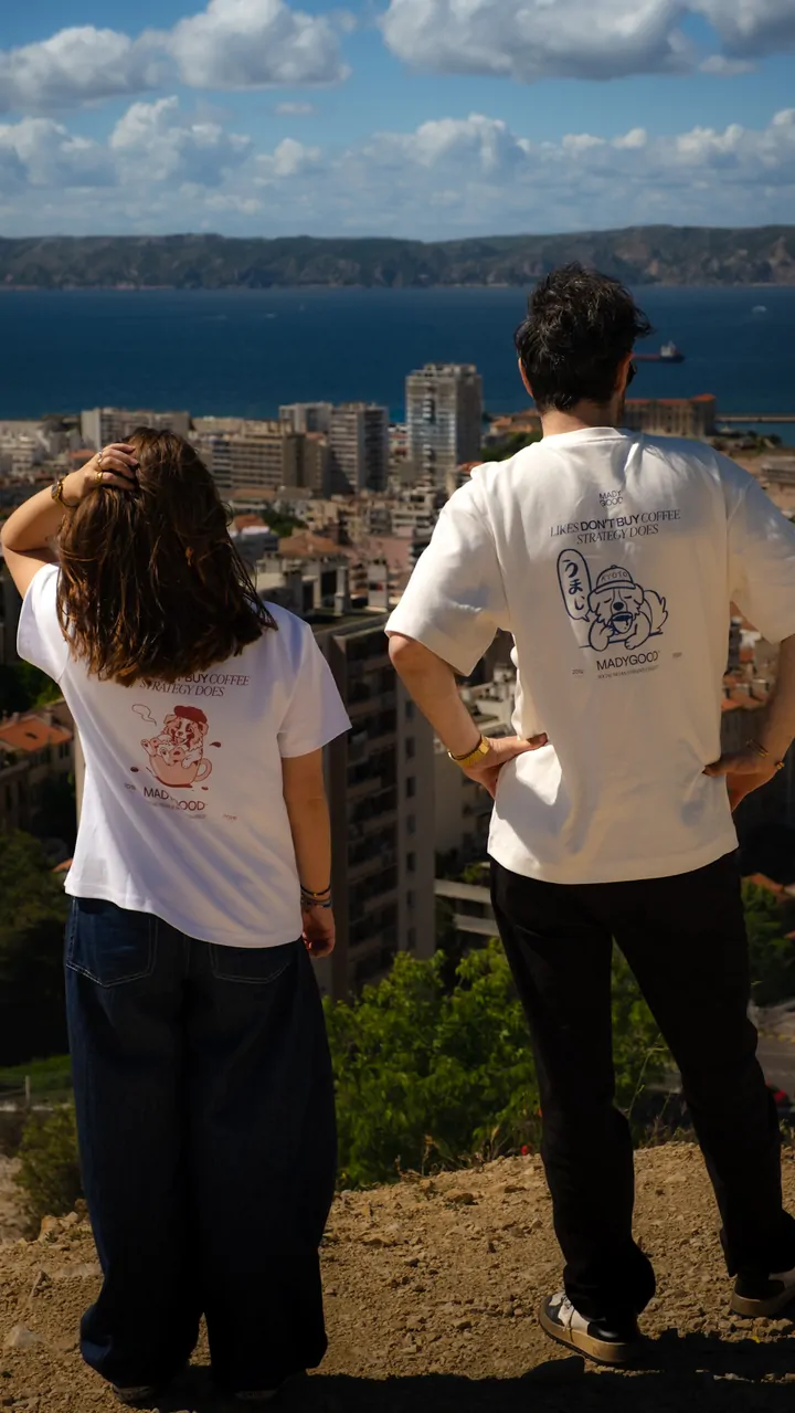 Deux personnes de dos, l’une en blanc, l’autre en écru, regardent la ville et la baie depuis la colline.