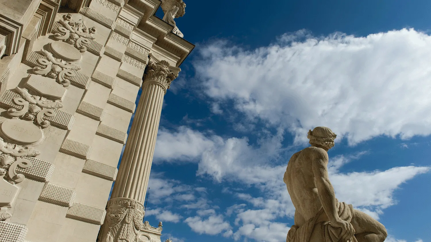 Une façade sculptée et une statue assise sous de grands nuages blancs.