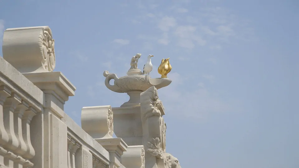 Une lampe antique en pierre et dorure posée sur une balustrade blanche, sous un ciel clair.